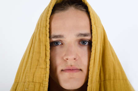 A portrait of the teenager boy covered with blanket looking at camera against white background. The young man wrapped himself in a warm blanket basks on a white background.の写真素材