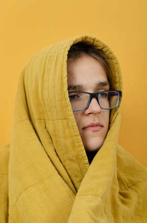 A portrait of the teenager boy in glasses covered with blanket against white background. The young man in glasses wrapped himself in a warm blanket basks on a white background.の写真素材
