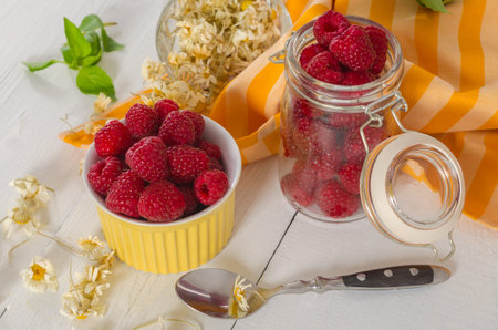 Raspberries in a bowl and a jar on the kitchen table with dry flowers and a spoon. Fresh food for a...