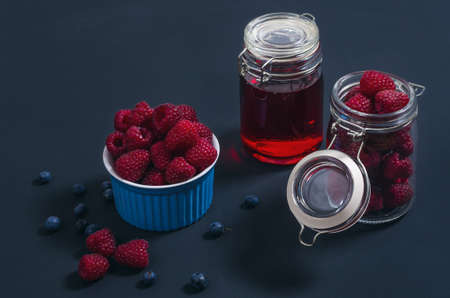 Two small jars with raspberries and syrup and berries in a bowl on a black stylish background. Fresh food for a healthy life and energy. Kitchen ingredients for home and family meals.の写真素材