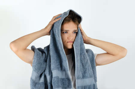 Cute caucasian teenager boy is drying hair with grey terry bath towel on white background. Young handsome man doing morning hygiene routine.の写真素材
