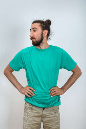 A bearded man who looking sideways with hands on hips. A young attractive man in a turquoise blue t-shirt poses for the camera. Studio portrait in casual clothes. Emotional, natural and sensual poses.の写真素材