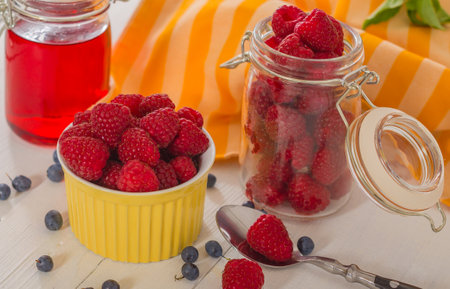 Raspberries in a bowl and a jar on the kitchen table with blueberries and a spoon. Fresh food for a healthy life and energy. Kitchen ingredients for home and family meals.の写真素材