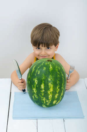 Cute child grinning holding a knife and fork near a watermelon. Juicy fruits or berries are very useful for children. Vitamins for babies, kids and teenagers at the white wooden table in studio.の写真素材