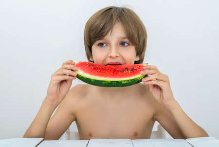 Hairy boy bites a watermelon and looks away. Juicy fruits or berries are very useful for children. Vitamins for babies, kids and teenagers at the white wooden table in studio.の写真素材