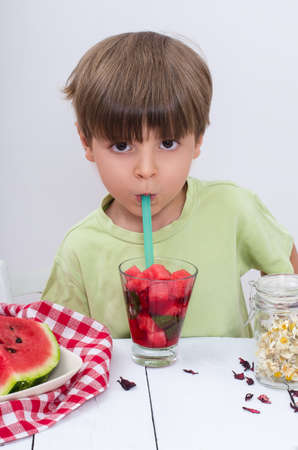The cute boy in light green t-shirt drinks watermelon cocktail through straw and looks straight. Useful berries and fruits for children of any age. Happy children drink vitamin smoothies in summer.の写真素材