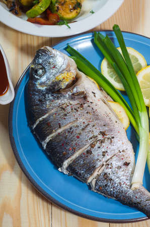 Baked fish on a blue plate with lemons and green onion at wooden table. Healthy seafood that gives a lot of energy at home. Delicious family breakfasts, lunches and dinners.の写真素材