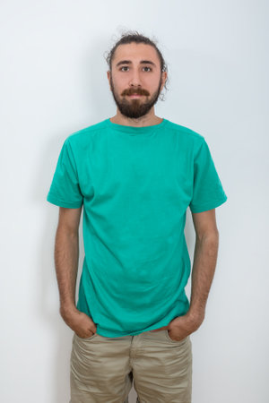 A bearded man put his hands in his pockets and looks ahead. A young attractive man in a turquoise blue t-shirt poses for the camera. Studio portrait in casual clothes. Emotional, natural and sensual poses.の写真素材