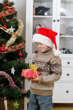 A cute boy in Santa's hat holding gift in hands near Christmas tree and looking at it. New Year's mood and festive magic near the Christmas tree create a cozy homely atmosphere.の写真素材