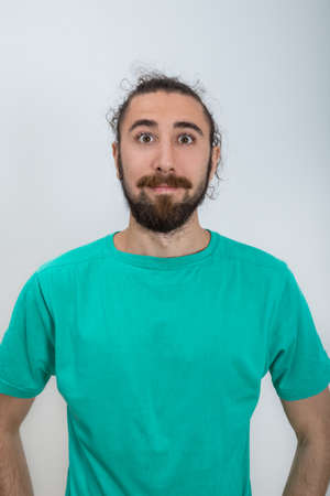 Surprised bearded man who looking straight with hands on hips. A young attractive man in a turquoise blue t-shirt poses for the camera. Studio portrait in casual clothes. Emotional, natural and sensual poses.の写真素材