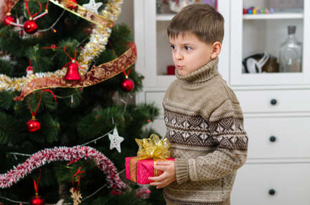A cute boy in brown winter sweater holding gift with bow in two hands and looking away. New Year's mood and festive magic near the Christmas tree create a cozy homely atmosphere.の写真素材