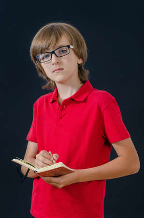 Cute boy making notes in a notebook and looking straight ahead. Smart and tidy child on training in a red t-shirt. Quality childhood education for serious kids.の写真素材