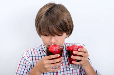 Teenage boy in a plaid shirt sniffs two glasses with pieces of watermelon. Useful berries and fruits for children of any age. Happy children drink vitamin smoothies in summer.の写真素材