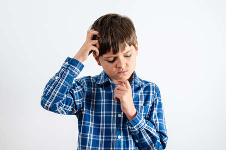 Caucasian boy in a blue plaid shirt is thinking about something on a white background. Portrait of a child with one hand in his hair and touches his chin.の写真素材