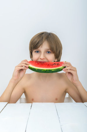 Hairy boy put watermelon in his mouth. Juicy fruits or berries are very useful for children. Vitamins for babies, kids and teenagers at the white wooden table in studio.の写真素材