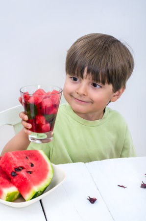 The cute boy in light green t-shirt holds watermelon cocktail with his hand and looks at it. Useful berries and fruits for children of any age. Happy children drink vitamin smoothies in summer.の写真素材