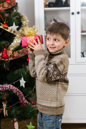 A cute boy in brown winter sweater put Christmas gift with bow to his ear and guess what's in it. New Year's mood and festive magic near the Christmas tree create a cozy homely atmosphere.の写真素材