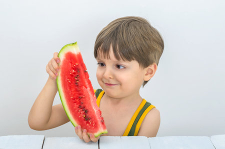 Cute child holding a watermelon and looking at it. Juicy fruits or berries are very useful for children. Vitamins for babies, kids and teenagers at the white wooden table in studio.の写真素材