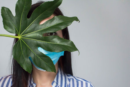 Beautiful woman hides face in a medical mask and open her eyes with big green tropical leaf, Facelessの写真素材