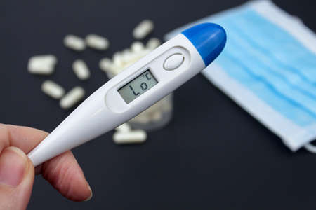 Hand holding white electronic thermometer in the background with packs of tablets and blue medical face mask. Selective focus.の写真素材