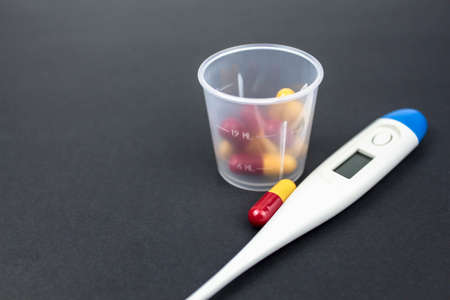 Medical capsules, tablets and clinical thermometer inside measuring cup on black background. Side viewの写真素材