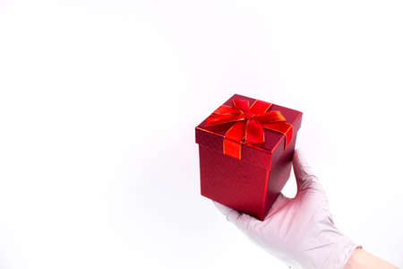 Women's hand in medical gloves hold a gift box with a red ribbon on a white background. Concept of sales and gifts during Coronavirus. Quarantine. Delivery courier. Online purchaseの写真素材