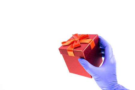 Women's hand in medical gloves hold a gift box with a red ribbon on a white background. Concept of sales and gifts during Coronavirus. Quarantine. Delivery courier. Online purchaseの写真素材