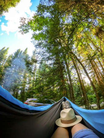 Woman relaxing in the hammock by the lake in the forest, POV view of legs in trekking boots with straw hat. Wanderlust concept sceneの写真素材