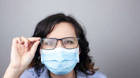 Young woman with foggy glasses caused by wearing disposable mask on white background. Protective measure during coronavirus pandemicの写真素材