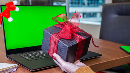 Hand holding big gift box next to laptop with green screen mockup, christmas shopping. Online shopping. New Year. Celebration, working from home officeの写真素材