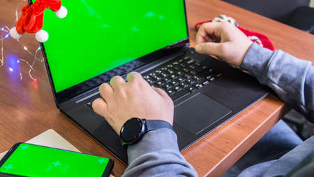 Closeup man hands typing on laptop keyboard with empty green screen, smartphone mockup and Christmas decoration on background. Holiday concept. Office. Mock-up.の写真素材