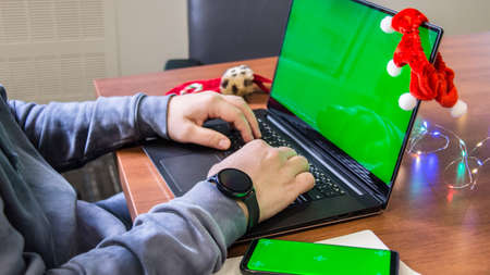 Closeup man hands typing on laptop keyboard with empty green screen, smartphone mockup and Christmas decoration on background. Holiday concept. Office. Mock-up.の写真素材