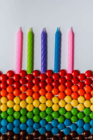 Rainbow of multicolored sweet candy dragees in shape of cake with candles on white background. Creative composition of round candy dragees in Rainbow colors. Concept birthday. Top view. Flat lay.の写真素材
