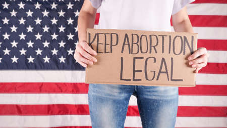 Young woman protester holds cardboard with Keep Abortion Legal sign against USA flag on background. Girl protesting against anti-abortion laws. Feminist power. Equal opportunity Womens rights reedom.の写真素材