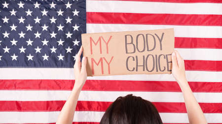 Young woman protester holds cardboard with My Body My Choice sign against USA flag on background. Girl protesting against anti-abortion laws. Feminist power. Equal opportunity Womens rights reedom.の写真素材