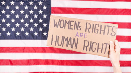 Woman is holding a sign Womens Rights Are Human Rights US flag on background. Protest against anti abortion law. Womens strike. Womens rights freedom.の写真素材