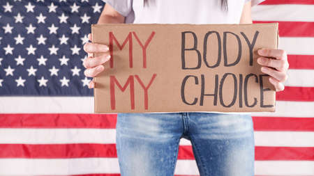 Young woman protester holds cardboard with My Body My Choice sign against USA flag on background. Girl protesting against anti-abortion laws. Feminist power. Equal opportunity Womens rights reedom.の写真素材