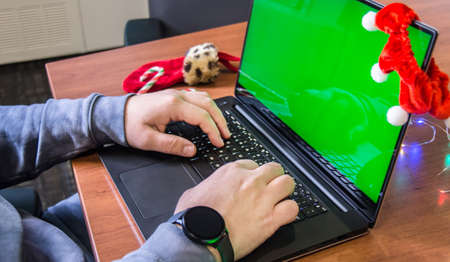 Closeup man hands typing on laptop keyboard with empty green screen and Christmas decoration on background. Holiday concept. Office. Mock-up.の写真素材