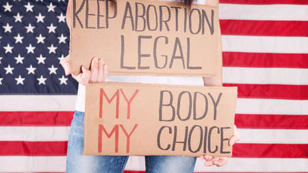 Young woman protester holds cardboard with Keep Abortion Legal and My Body My Choice signs against USA flag on background. Girl protesting against anti-abortion laws. Womens rights freedom.の写真素材