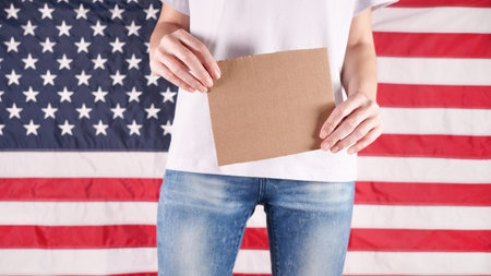 Unrecognizable woman showing empty cardboard banner. Hands holding blank poster from a cardboard box. Space for your slogan, logo or advertisement. Banner design concept. American flag on background.の写真素材
