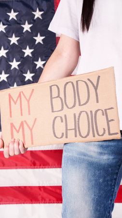 Young woman protester holds cardboard with My Body My Choice sign against USA flag on background. Girl protesting against anti-abortion laws. Feminist power. Equal opportunity Womens rights reedomの写真素材