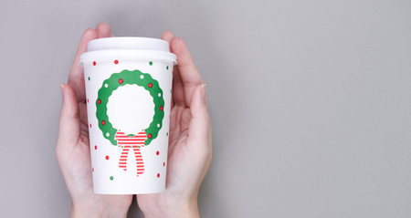 Hands holding craft paper coffee cup, grey background. Christmas or New Year holiday greeting paper...