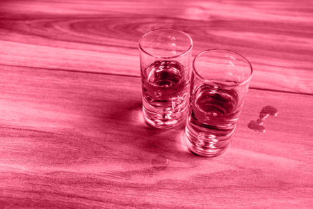 Two shot glasses with whiskey on a wooden table. Image tonned with Viva Magenta color of year 2023の写真素材