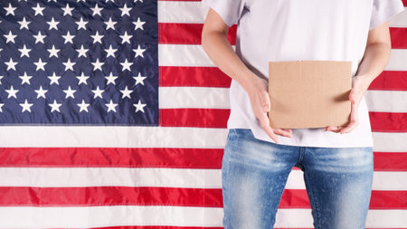Unrecognizable woman is holding an empty cardboard with Space for Text sign US flag on background. Vote right. Protest against law. Womens rights freedom. Against racism racial discrimination.の写真素材