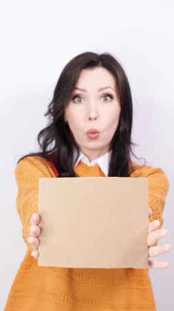 Beautiful young surprized brunette woman in yellow sweater holding blank placard with Copy Space for advertisement. Selective focus, vertical. Empty billboard with space for text white backgroundの写真素材