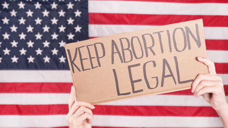 Young woman protester holds cardboard with Keep Abortion Legal sign against USA flag on background. Girl protesting against anti-abortion laws. Feminist power. Equal opportunity Womens rights reedomの写真素材