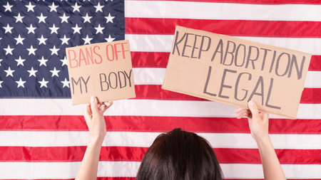 Young woman protester holds cardboard with Keep Abortion Legal and Bans Of My Body signs against USA flag on background. Girl protesting against anti-abortion laws. Womens rights freedomの写真素材