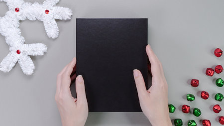 Template composition hands holding blank black box, gift box, product packaging on grey background with holiday decore. Mockup, photo card, Christmas card, greeting card.の写真素材