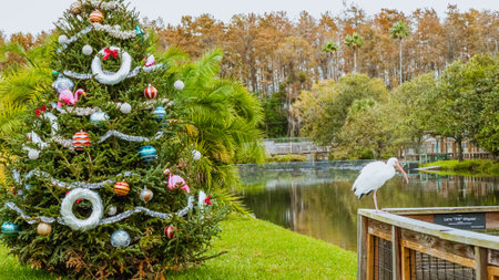 Tree adorned with colorful ornaments, lights, star. Serene lake surrounded by lush green trees and shrubs. White bird perched on branch by waters edge. Cloudy sky with hints of blue.の写真素材