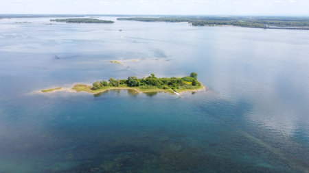 Aerial view of a small green island in a large blue water body on a partly cloudy day. The island is lush with trees, surrounded by calm water, with a clear blue sky above.の写真素材